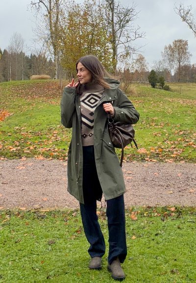 Mujer con abrigo verde y suéter estampado sosteniendo una bolsa marrón, de pie sobre la hierba cerca de un camino con árboles otoñales de fondo.