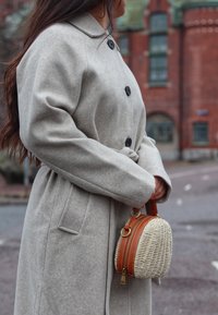 Light grey wool coat with a collar, featuring large black buttons and a tie waist. Holding a round woven handbag with leather accents.