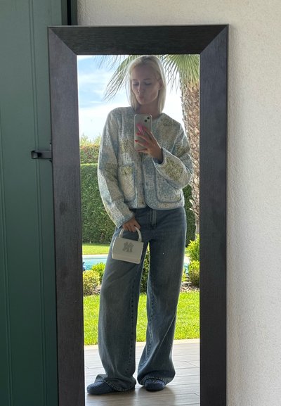 Chaqueta acolchada azul con flores y bolsillos, jeans de mezclilla holgados y un bolso blanco con logo. De pie frente a un espejo.