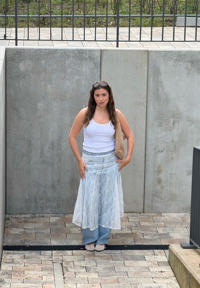 Mujer con cabello largo y oscuro está apoyada contra una pared de hormigón, lleva una camiseta blanca sin mangas, falda de encaje en capas sobre vaqueros, gafas en la cabeza y bolso beige al hombro.