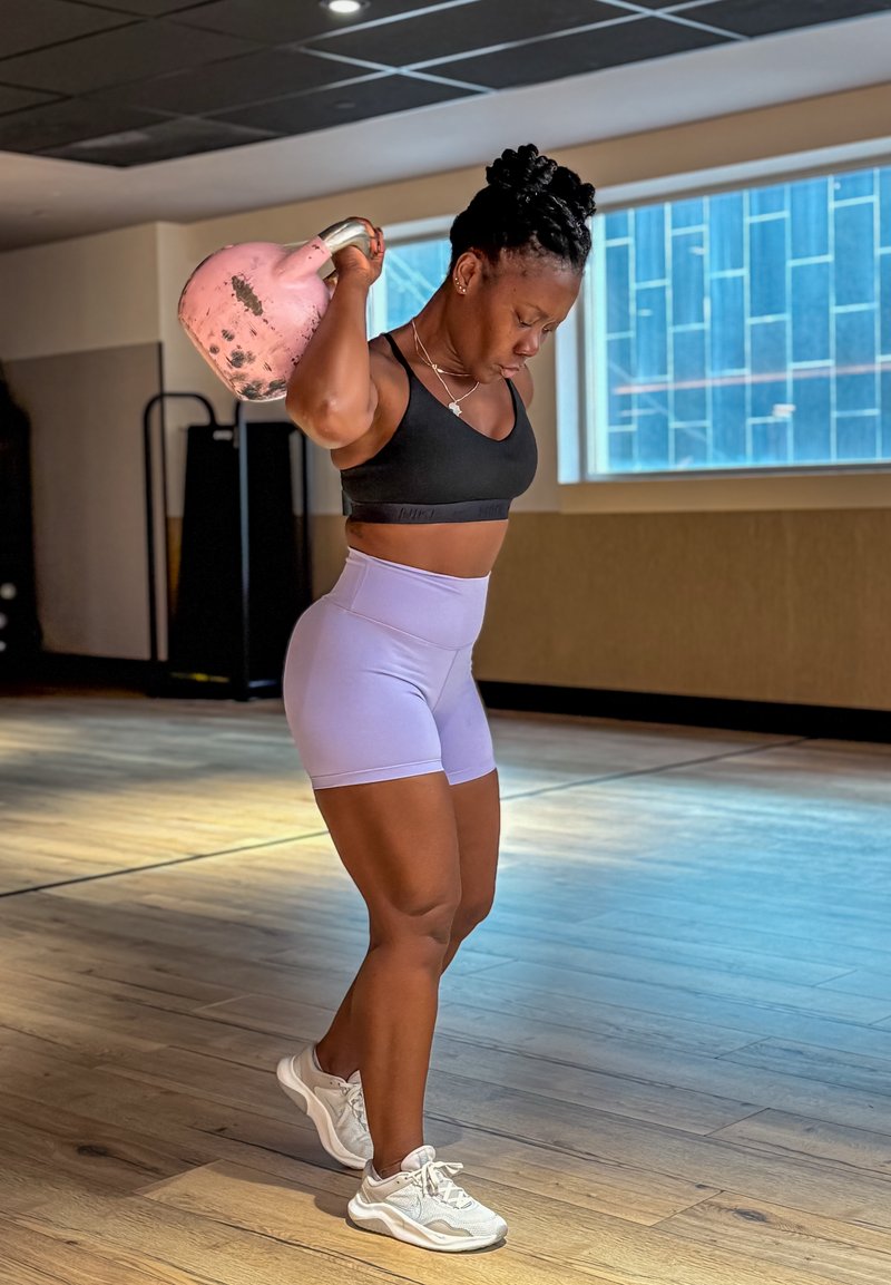Una persona solleva un kettlebell rosa, indossando un reggiseno sportivo nero e pantaloni corti lavanda. Il pavimento è in legno e ci sono elementi da palestra sullo sfondo.