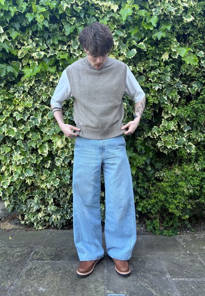 Chaleco beige de punto sobre una camisa de manga corta blanca, combinado con jeans de pierna ancha de color azul claro y zapatos de cuero marrón, frente a un fondo lleno de hojas.