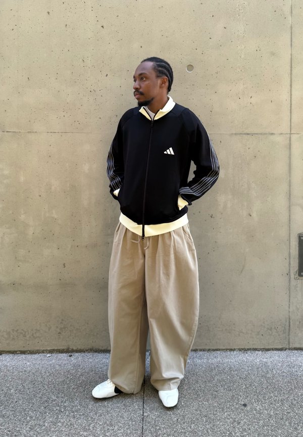 Homme aux cheveux tressés portant une veste noire zippée, un pantalon beige large, des chaussures blanches, debout les mains dans les poches contre un mur en béton.