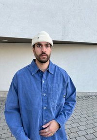 Camisa azul clara, com botões e um bolso frontal, combinada com um gorro de malha em creme. Textura lisa, corte largo, usada de forma casual.