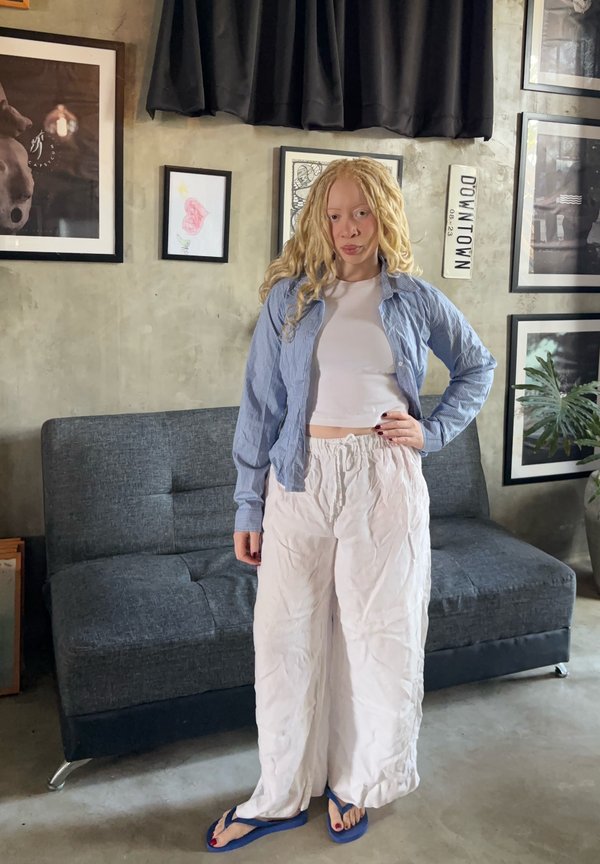 Giovane donna con lunghi capelli biondi ricci in piedi con la mano sui fianchi, indossa pantaloni bianchi, top corto bianco, camicia blu e infradito blu all’interno.