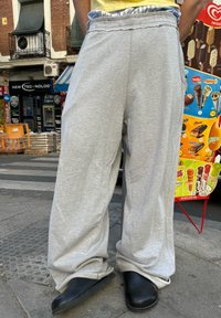 Világosszürke melegítőnadrág, rugalmas derékpánttal, laza szabású. Fekete slip-on cipőkkel párosítva, strukturált járólap háttérrel.