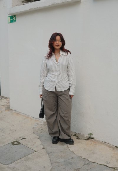 Camisa de botones a rayas en blanco y gris; pantalones grises de corte holgado; zapatos negros; collares en capas; bolso negro pequeño. Fondo liso.