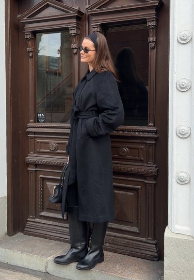 Mujer con abrigo negro, botas, gafas de sol y diadema, sostiene un bolso negro frente a una puerta de madera oscura y ornamentada.