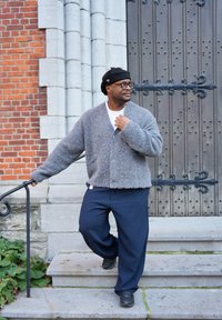 Cardigan en laine grise à la texture douce, boutonné à l'avant, porté sur une chemise blanche. Pantalon large de couleur foncée, chaussures noires et un bonnet noir.