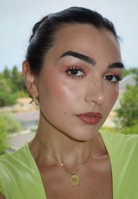 Pendientes en forma de estrella dorada, collar dorado con colgante circular, maquillaje natural en tonos cálidos y una blusa verde brillante. Iluminación suave.