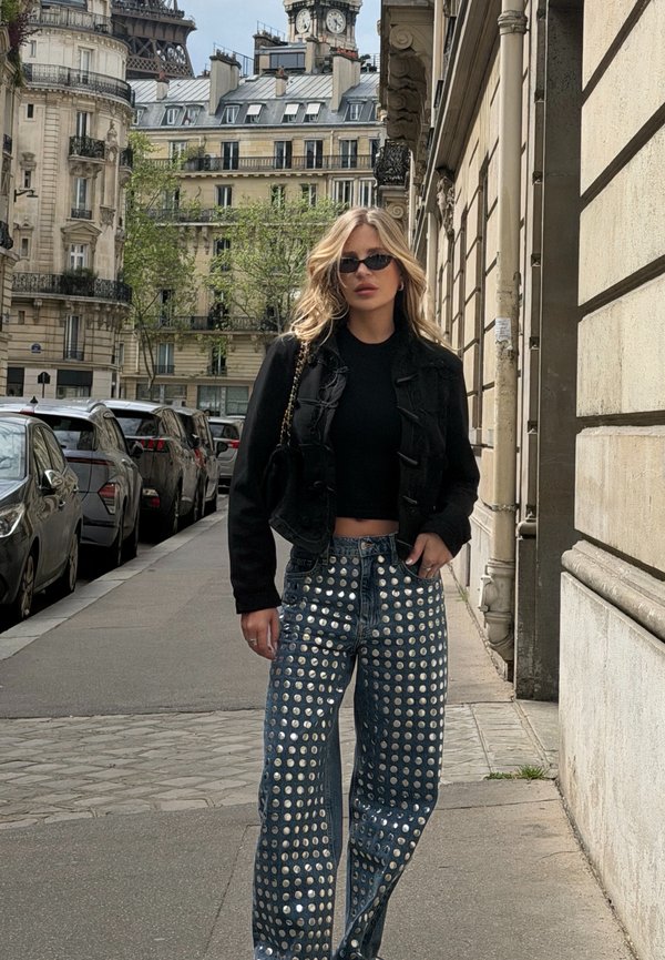 Femme blonde portant une veste courte noire, des lunettes de soleil et un jean bleu avec des clous circulaires argentés marchant dans une rue de la ville de Paris.