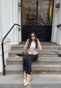 Chemise en satin argentée avec boutons, pantalon en satin noir, talons pointus crème. Assis sur des marches en pierre, avec une pochette métallique à proximité.