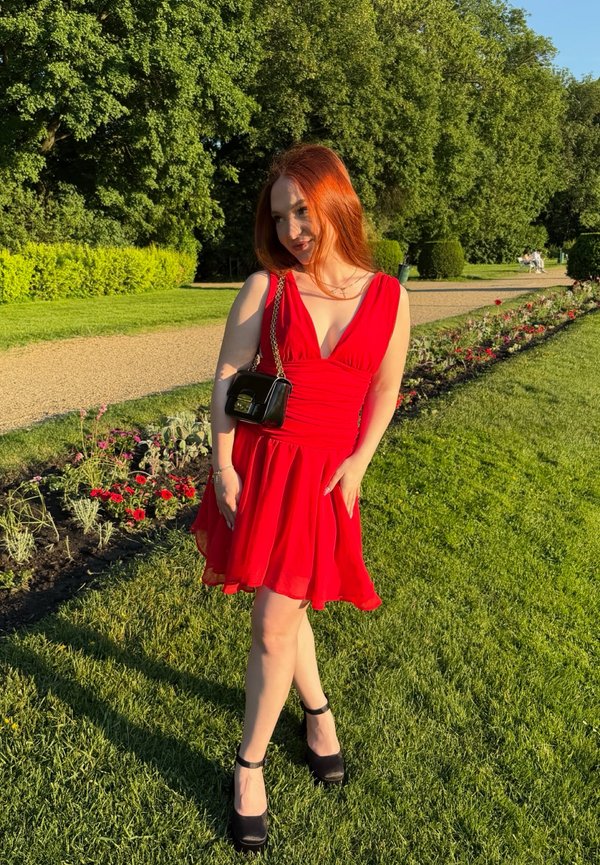 Jeune femme aux cheveux roux, vêtue d'une robe rouge et de chaussures noires, debout sur l'herbe près d'une plate-bande dans un parc ensoleillé.