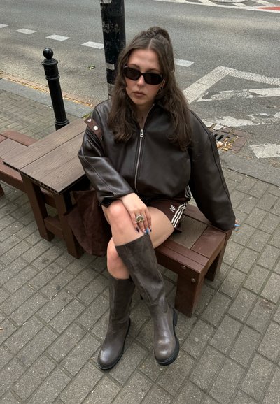 Chaqueta bomber de cuero marrón con herrajes plateados, gafas de sol negras, botas marrones hasta la rodilla y asientos exteriores marrones sobre una superficie pavimentada.