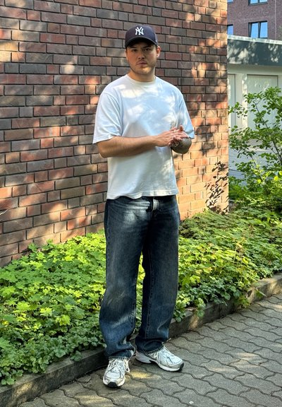 Camisa blanca de manga corta, vaqueros azules de mezclilla, zapatillas grises y una gorra de béisbol oscura con un logo, de pie contra una pared de ladrillos y vegetación.