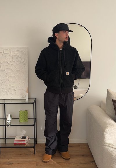 Chaqueta de forro polar negra con capucha, cierre frontal y bolsillos laterales. Combinada con pantalones negros y botas marrones. De pie junto a un espejo y una estantería.