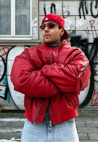 Homme portant une veste en cuir rouge, une casquette rouge à l'envers et des lunettes de soleil, debout les bras croisés devant un mur couvert de graffitis.