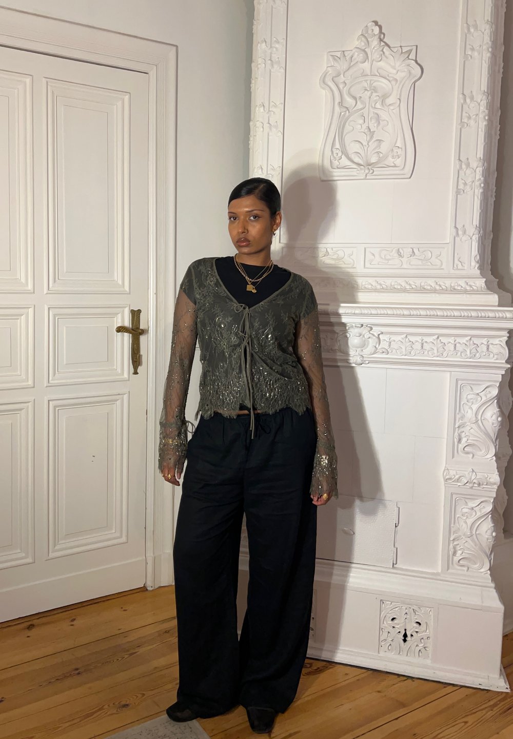 Olivi vihreä pitsinen crop-top, jossa on solmittava suljin, pitkät läpinäkyvät hihat ja yksityiskohtainen muotoilu, yhdistettynä löysiin mustiin korkeavyötäröisiin housuihin.