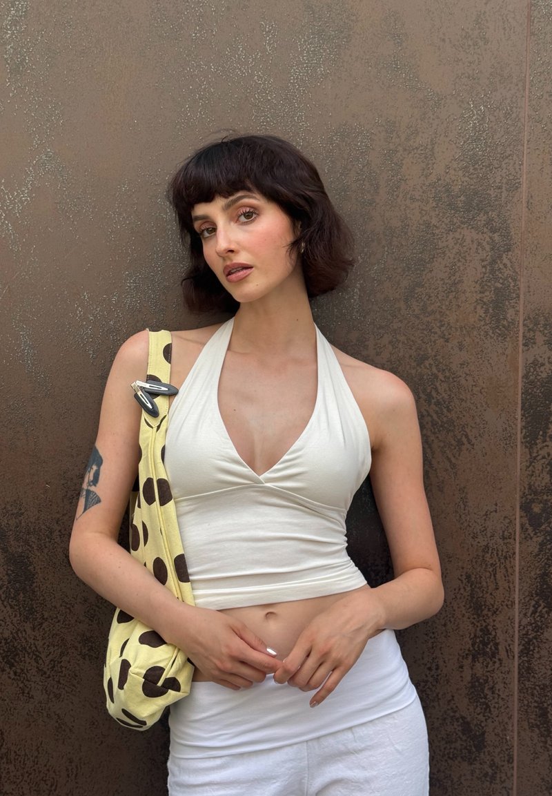 Femme aux cheveux courts et foncés se tenant contre un mur brun texturé, portant un haut dos nu blanc, un pantalon blanc et un sac à main jaune à pois avec des clips.