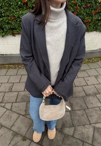Femme portant un blazer gris, un pull à col roulé crème, un jean bleu, des bottes marron clair, tenant un petit sac à main beige en daim, debout sur un trottoir pavé.