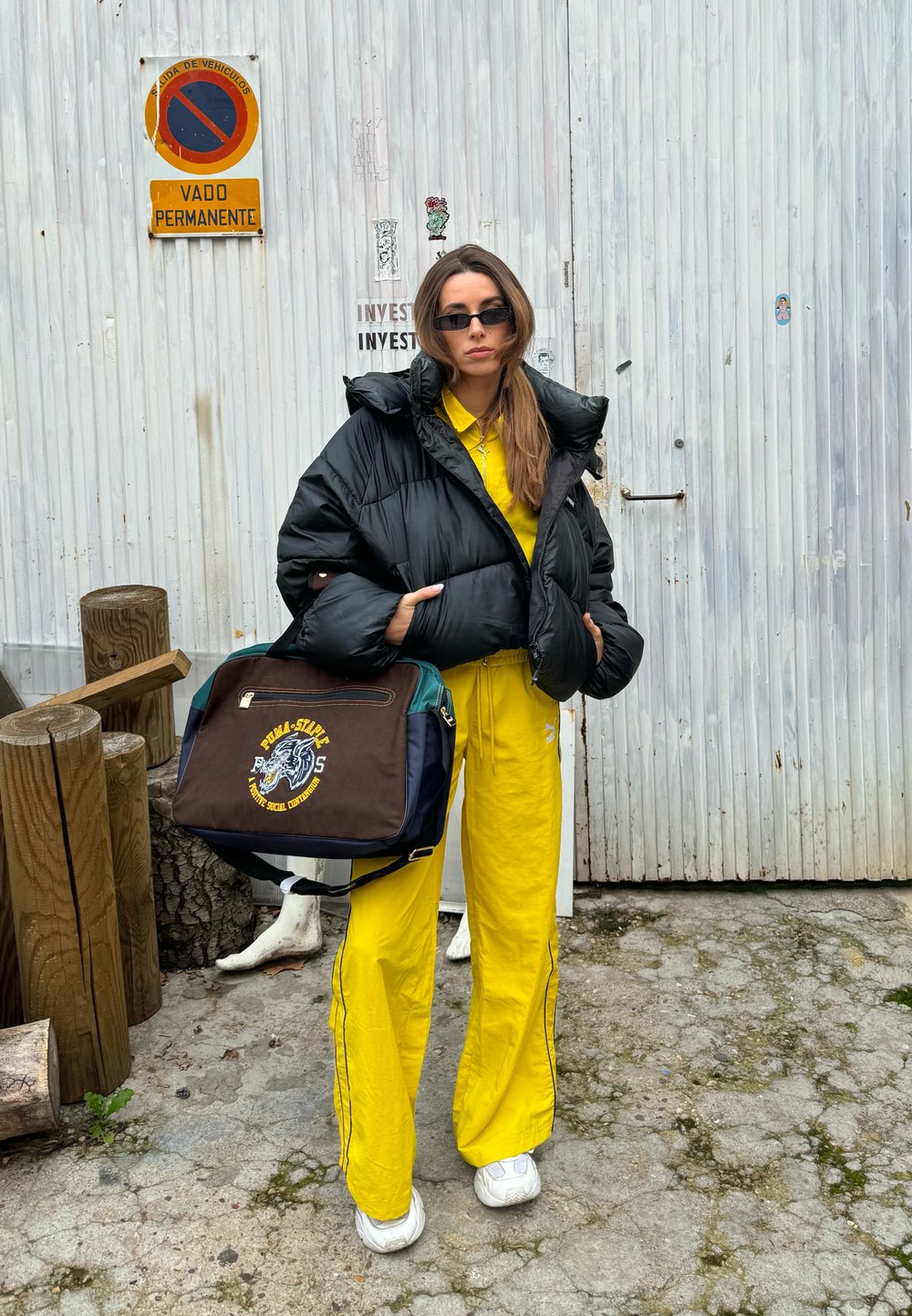 Μαύρο φουσκωτό μπουφάν, κίτρινο κοστούμι προπόνησης και λευκά αθλητικά παπούτσια. Κρατά μια πράσινη και καφέ τσάντα με κεντητό σχέδιο. Ξύλινο και πέτρινο φόντο.