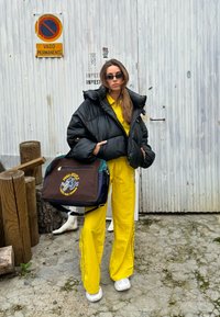 Μαύρο φουσκωτό μπουφάν, κίτρινο κοστούμι προπόνησης και λευκά αθλητικά παπούτσια. Κρατά μια πράσινη και καφέ τσάντα με κεντητό σχέδιο. Ξύλινο και πέτρινο φόντο.