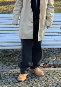 Osoba v béžovom kabáte, čiernych nohaviciach a hnedých topánkach stojaca na kockovanej dlažbe pred bielou lavičkou vonku.