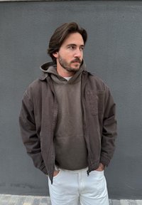 Man with brown hair and beard wearing a brown jacket and hoodie, standing with hands in pockets against a grey wall.