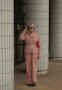 Rosa skreddersydd kostyme med vide bukser, kjedebelte og rød håndveske. Tilbehøret inkluderer solbriller og lagdelte halskjeder.