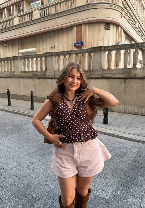 Ung kvinna med långt hår som bär en prickig topp, ljusa shorts och bruna stövlar, poserar på en stadstrottoar framför en stenbyggnad.