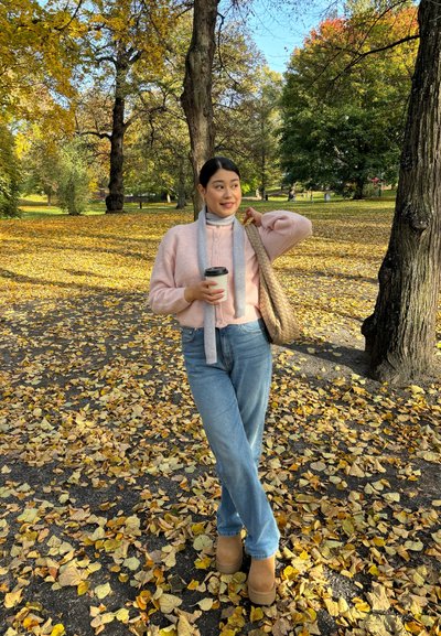 Suéter de punto rosa claro, bufanda gris, jeans azules y botas marrones. Sostenido una taza para llevar, de pie sobre un suelo cubierto de hojas amarillas.