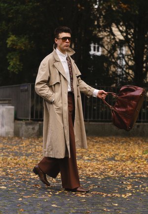 Beige trench coat over a white shirt, brown trousers, and leather shoes. Holding a dark brown bag, walking on leaf-strewn pavement.