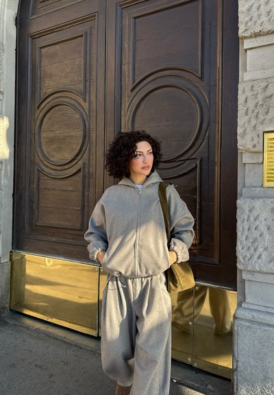 Mujer con cabello rizado, vestida con sudadera y pantalones grises, de pie con las manos en los bolsillos junto a una gran puerta de madera con paneles de latón.