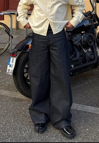 Széles szárú sötétkék farmernadrág magas derékkal, világos színű gombos inggel és fényes fekete cipővel párosítva.