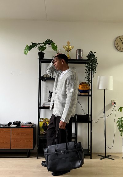 Hombre con cárdigan gris y pantalones negros sostiene una bolsa negra, de pie cerca de una estantería negra con plantas, un trofeo y un balón de baloncesto.