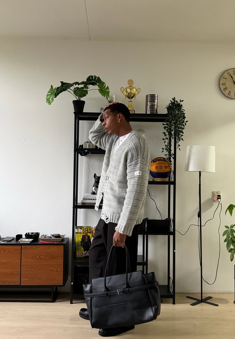 Un homme portant un cardigan gris et un pantalon noir tient un sac noir, debout près d'une étagère noire avec des plantes, un trophée et un ballon de basket.