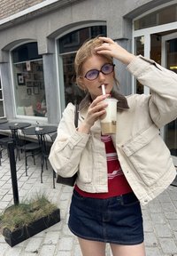 Lys beige lerretjakke med brun kordfløyelskrage, rødstripet genser under og mørk denimskjørt; holder en drink med sugerør.