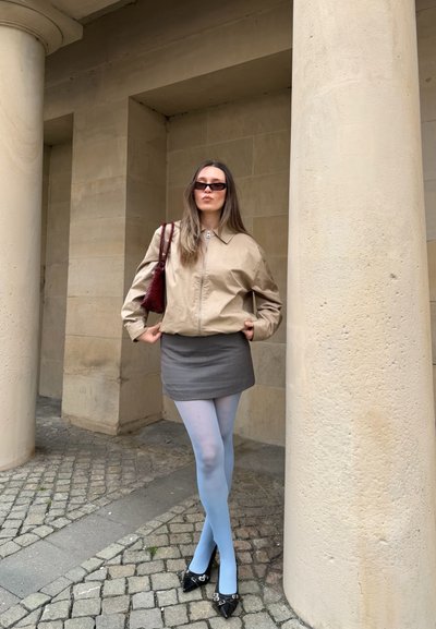 Mujer con chaqueta beige, minifalda gris, medias azules claras, zapatos negros puntiagudos y gafas de sol oscuras, está de pie entre grandes columnas de piedra.