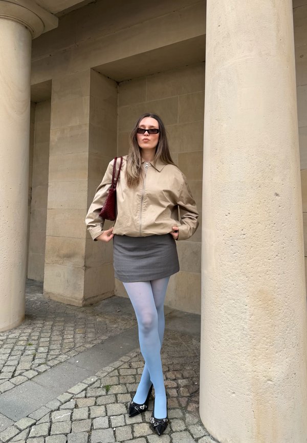 Femme portant une veste beige, une mini-jupe grise, des collants bleu clair, des chaussures noires pointues et des lunettes de soleil foncées, debout entre de grandes colonnes en pierre.