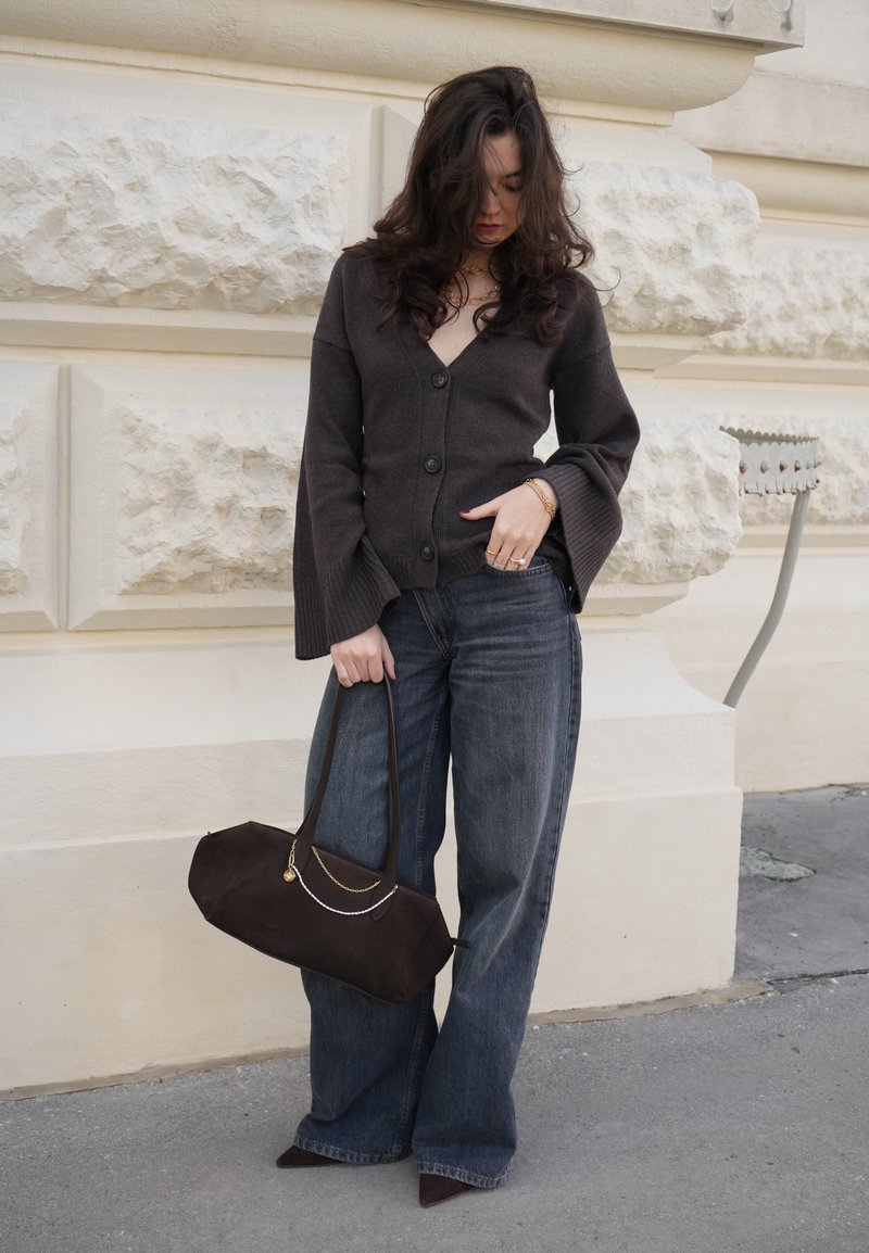 Femme aux longs cheveux foncés portant un cardigan marron foncé et un jean à jambes larges, tenant un grand sac à main sombre, debout près d'un mur en pierre texturé.