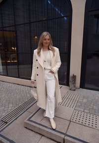 Casaco longo creme com botões pretos, suéter creme justo, calças brancas, mocassins creme e uma bolsa pequena, com um fundo de edifício moderno.