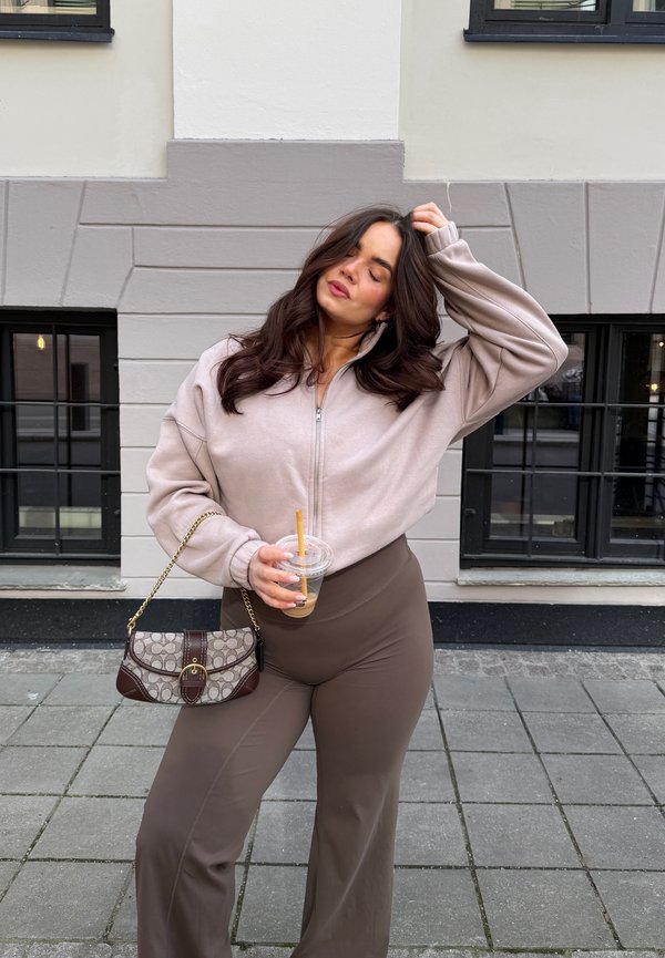 Femme aux longs cheveux foncés portant une veste beige zippée et un pantalon marron, tenant un café glacé et un petit sac à main à motifs, debout sur un trottoir en ville.