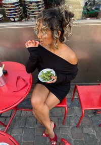 Svart off-shoulder kjole, røde sandaler, holder en tallerken med grøn edamame på et rødt bord. Stabler med dekorative skåler i bakgrunnen.