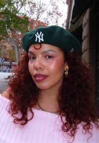 Jeune femme aux cheveux bouclés portant un béret vert foncé avec un logo NY blanc, des boucles d'oreilles dorées en anneau, un haut rose et un collier doré, en plein air avec des arbres en arrière-plan.