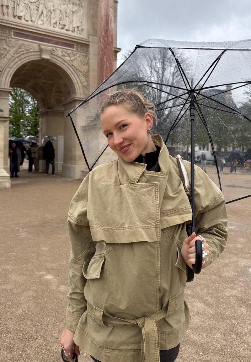 Femme en veste beige tenant un parapluie transparent par une journée pluvieuse près d'une arche en pierre ornée avec des personnes en arrière-plan.