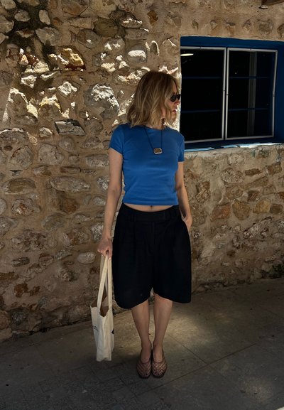 Mujer con top azul corto y pantalones cortos negros está de pie junto a una pared de piedra, sosteniendo una bolsa de tela y mirando hacia un lado cerca de una ventana.