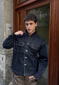 Veste en denim foncé avec un col structuré, des poches avant et des coutures contrastantes. Portée sur un pantalon clair. Couleur unie, texture lisse.