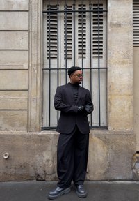 Veste de costume noire par-dessus un col roulé noir, associée à un pantalon large noir et des gants. Chaussures grises. Sur un fond de mur en pierre texturé.