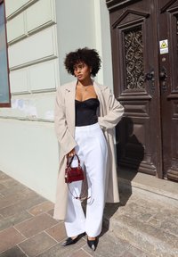 Casaco oversized bege claro sobre um top de corpete preto, combinado com calças brancas de perna larga. Os acessórios incluem uma pequena mala vermelha e sapatos pretos de bico.
