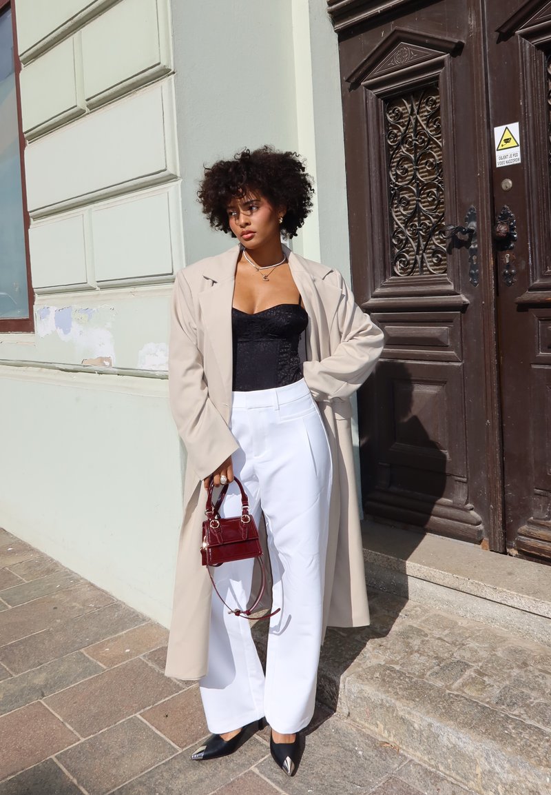 Light beige oversized coat over a black corset top, paired with white wide-leg trousers. Accessories include a small red handbag and black pointed shoes.